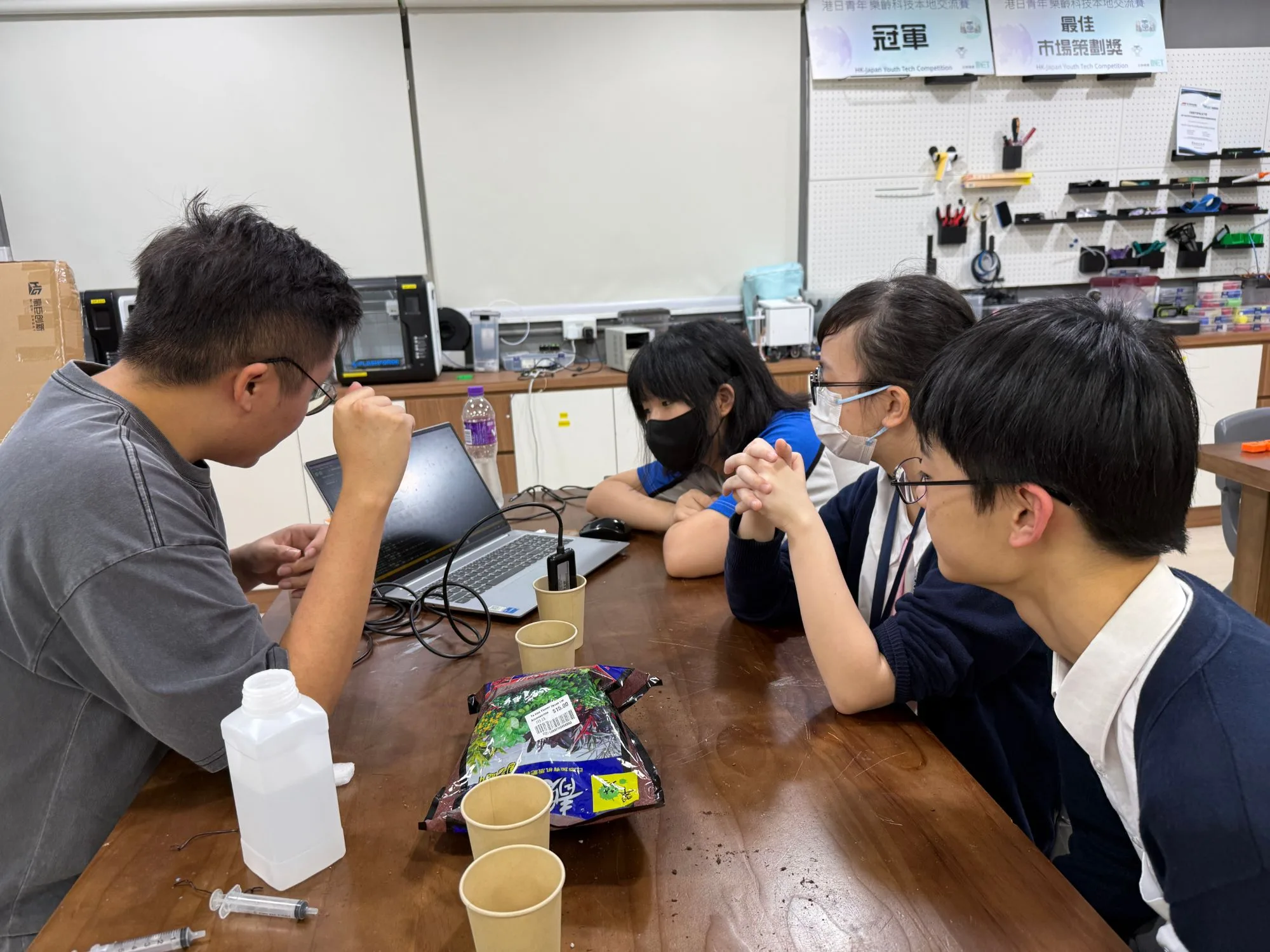 A project-based teaching scene in a STEM studio: culture reagents, syringes and electronic parts lie on the table while an instructor guides students in using a computer to set up and test an IoT project.