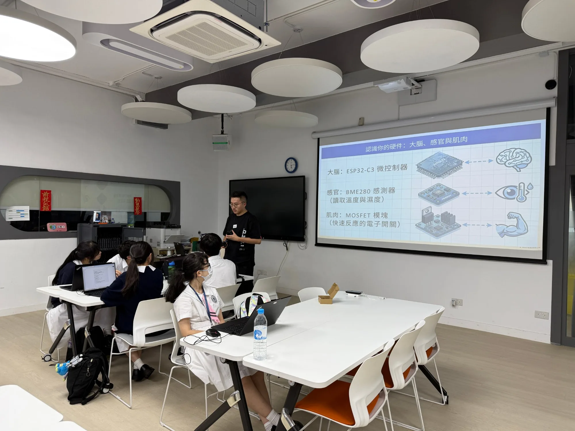 A teaching session on IoT hardware architecture: the slide uses a brain, senses and muscles as analogies for the microcontroller, the temperature and humidity sensor and the electronic switch module, while students seated with laptops listen and learn.