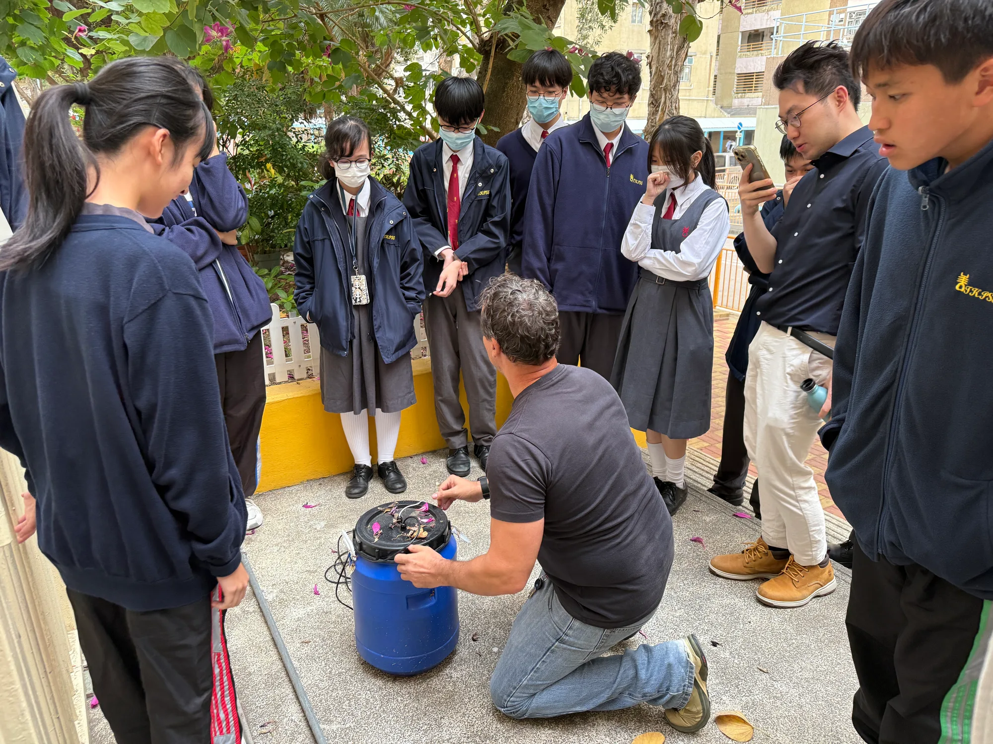 校園戶外實作活動，導師蹲在藍色收集桶旁向學生示範處理果皮等材料，一群穿校服的學生圍著觀看。