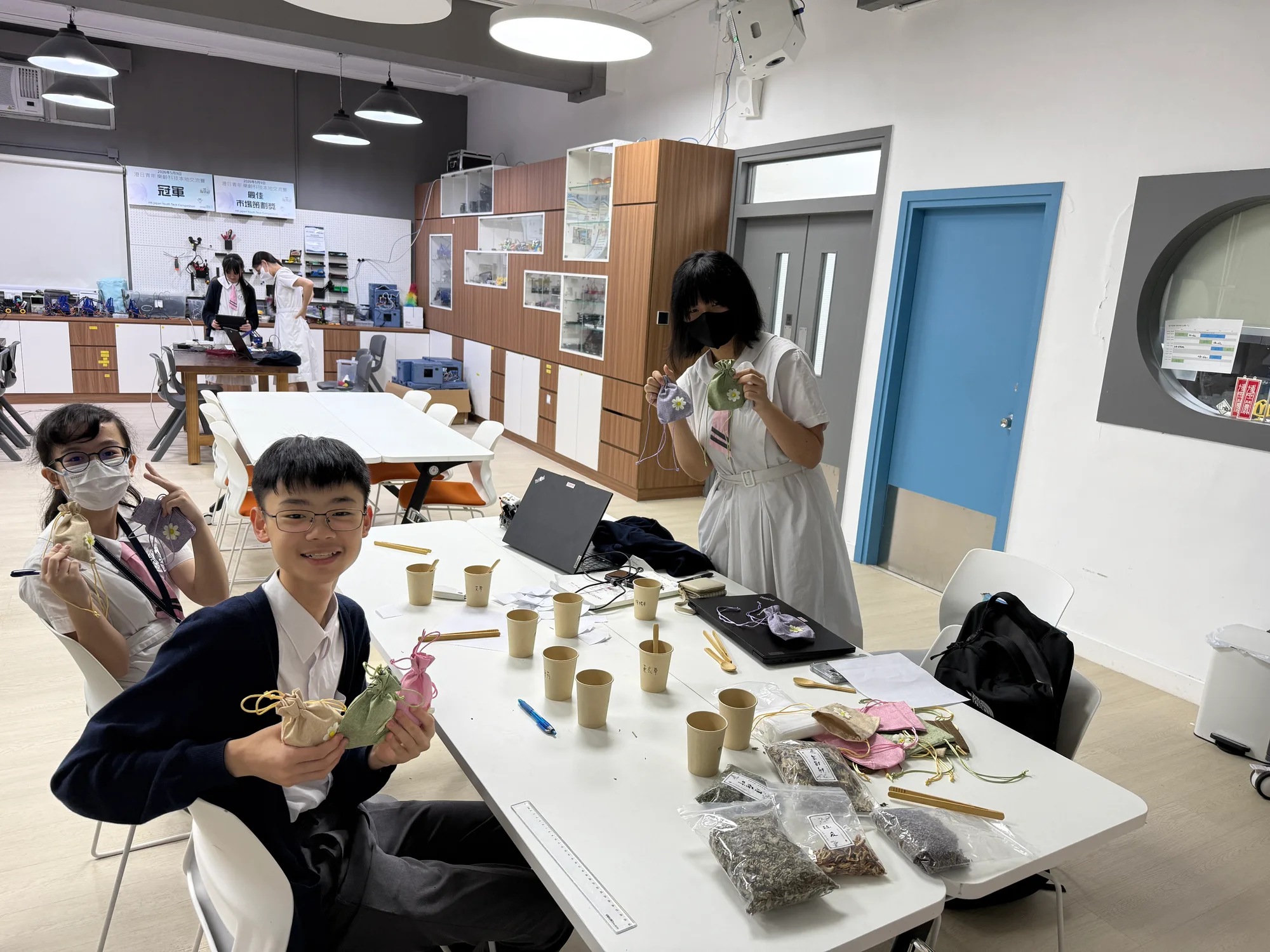 學校活動室內，幾名學生圍著長枱製作香包，枱面放有紙杯、麻繩和乾花材料，數名學生舉起完成的麻布香包展示。
