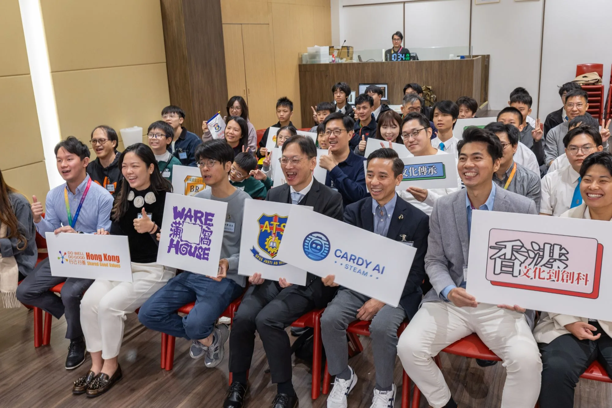 Group photo at a technology and culture event: guests in the front row hold signs such as "From Hong Kong Culture to Innovation and Technology", "CARDY AI STEAM" and "WARE HOUSE", while the seats behind are filled with students and instructors taking part in the project, recording the moment of results sharing and exchange.