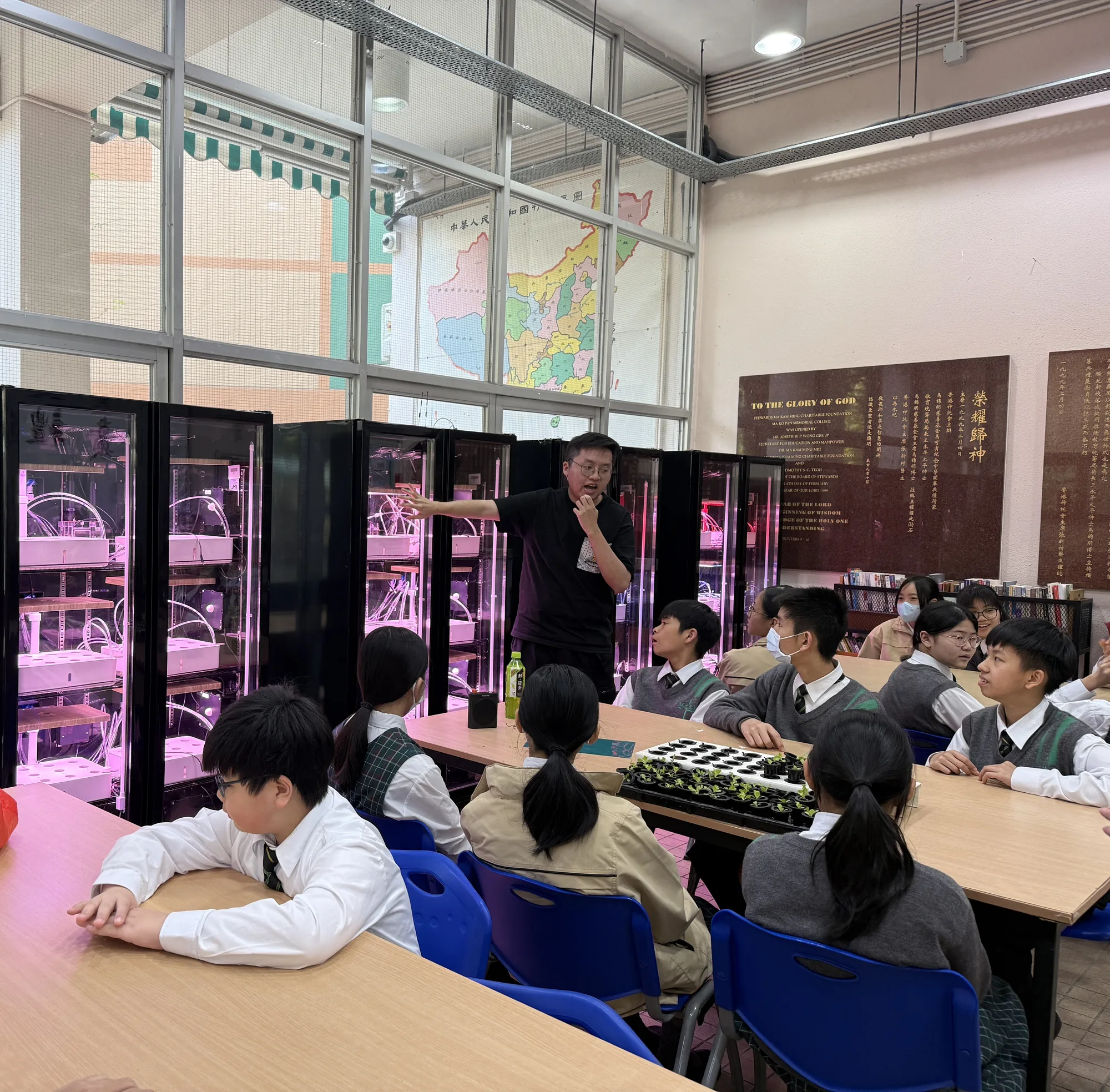 An instructor explains to students indoors how a vertical smart hydroponic growing cabinet works; seedling trays sit on the table and the students sit around listening.